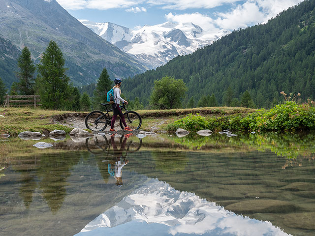 woman on mountain bike passing by the edge of an alpine lake with glacier view